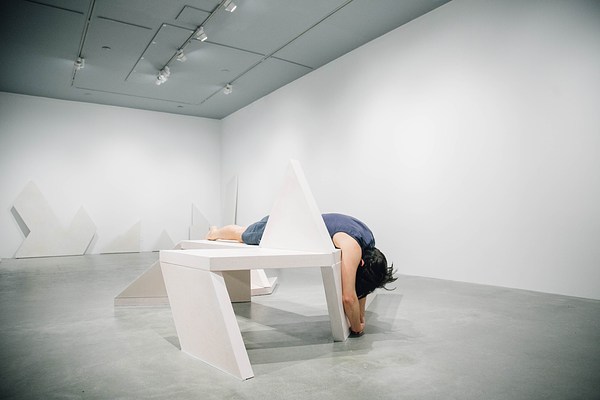 A performance piece in a gallery where a person wearing navy sits on a bench sculpture with his head folded into his lap and his arms towards the ground.