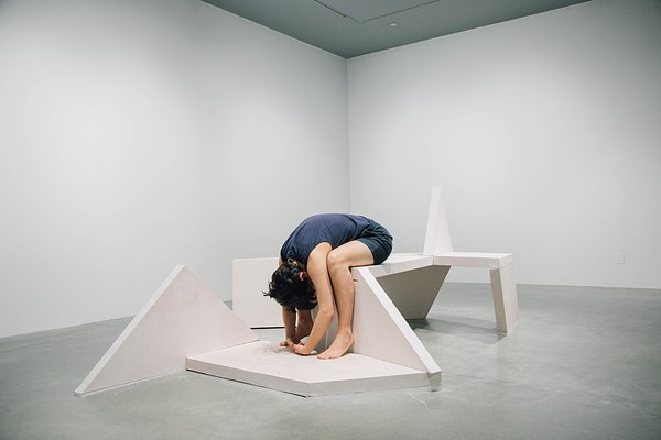 Close up of a performance piece in a gallery where a person wearing navy sits on a bench sculpture with his head folded into his lap and his arms towards the ground.