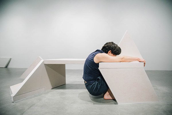 A performance piece where a person lies on a bench sculpture face down with their legs up in the air and their arms dragging on the ground.&nbsp;