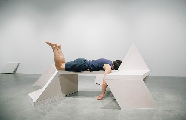 A performance piece where a person is on their knees golding their upper body up with their arms on a sculpture bench.