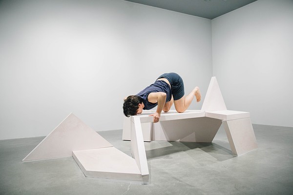 A performance piece where a person is hunched over beside a sculpture bench with their head resting low on the bench and their arms gripped behind their back.