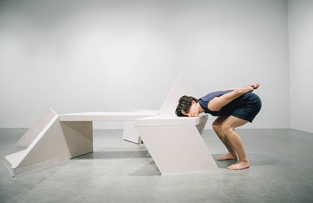 A performance piece where an older woman wearing a blue cardigan sits on a sculpture bench with her hands on her thighs.