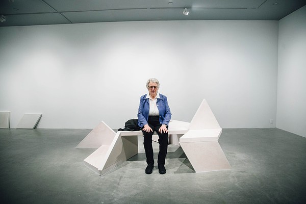 View from the side of a performance piece where an older woman wearing a blue cardigan sits on a sculpture bench with her hands on her thighs.