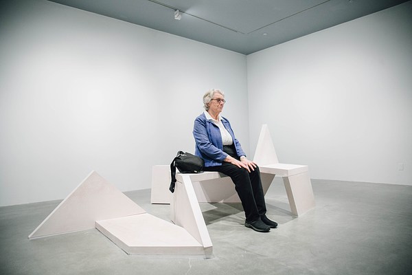 A performance piece in the gallery where a man sits up tall on a sculpture bench with his back to the camera.