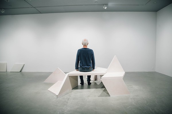 A performance piece in the gallery where two men sit up tall on a sculpture bench facing opposite directions.