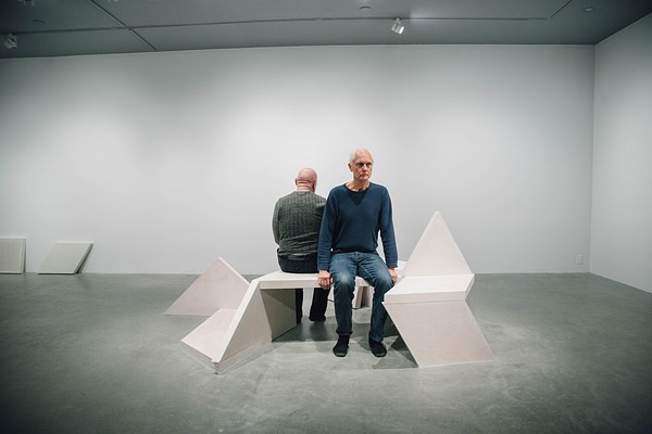 A performance piece in the gallery where a man in green sits facing the back right wall of the gallery on a sculpture bench and a man in blue stands facing the opposite wall with the bench behind him.