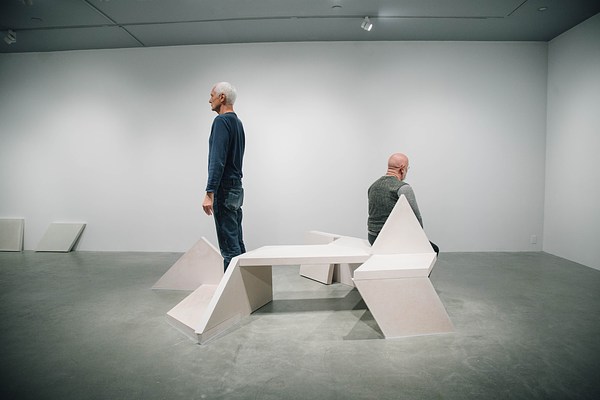 Performance piece where a man in green sits facing the right gallery wall and a man behind him stands up facing the opposite wall with a granite bench in between them.