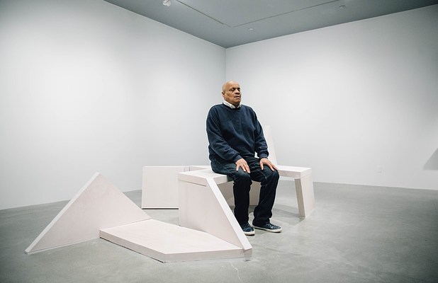 View from the side of a man sitting up tall on a sculpture bench as part of a performance piece.