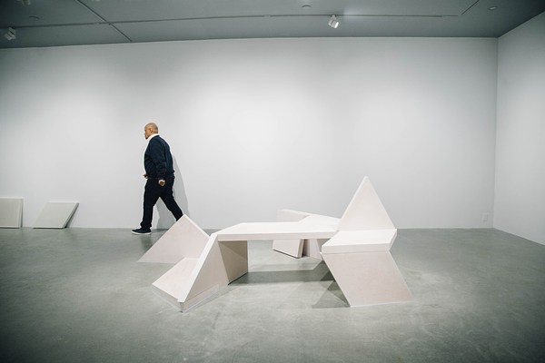 A concrete bench sculpture, made of geometric shapes with a triangle sticking up from the seat area, sits on a gray floor with a man walking against the back wall to the left in the background.
