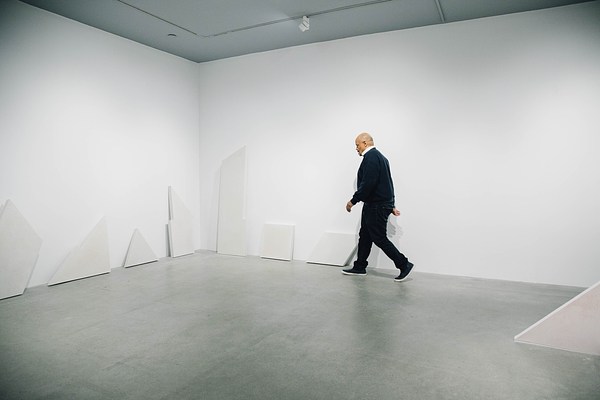 A gallery with geometric white shapes lining the walls and a man walking against the back wall to the left in the background.