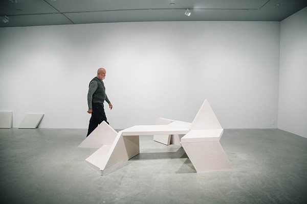 A concrete bench sculpture, made of geometric shapes with a triangle sticking up from the seat area and a man wearing green in the background walking from the left side of the gallery towards the right.