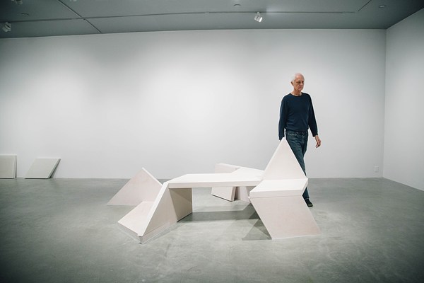A concrete bench sculpture, made of geometric shapes with a triangle sticking up from the seat area and a man in dark blue walking around the perimeter of the bench.