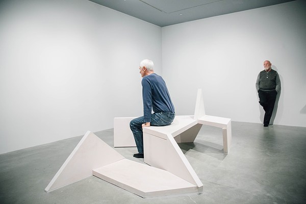 Performance piece where a man in green stands against a gallery wall facing a bench sculpture where a man in blue is sitting, facing the back wall of the gallery.