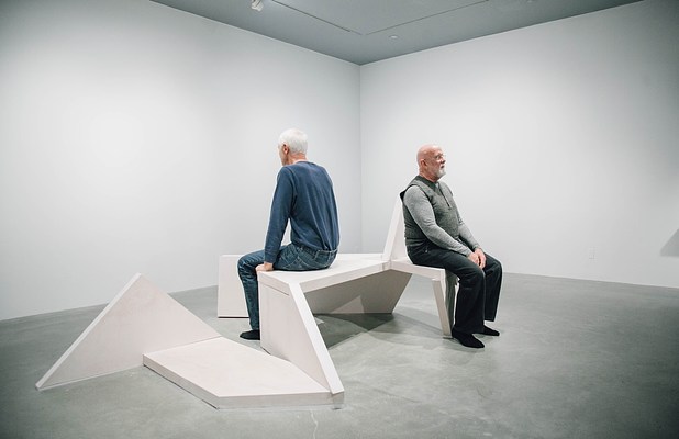 Performance piece where two men sit opposite one another facing away from each other on a sculpture bench in a gallery.. . 