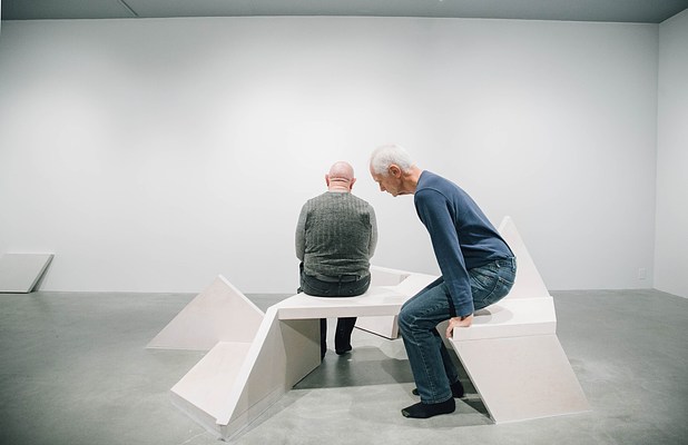 Performance piece where a man in green sits on a sculpture bench facing the back gallery wall and a man in blue goes to sit down near him on a different end of the bench.