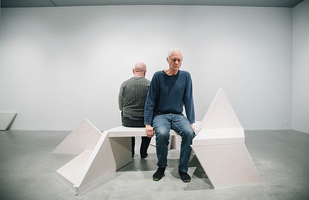 Two men sitting on opposite sides of a sculpture bench face away from each other.