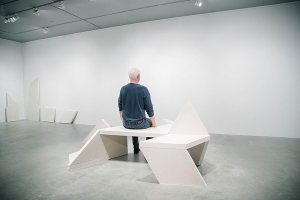A man in blue sits up straight on a sculpture bench facing the back wall and his back to the camera.