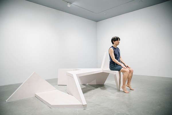 Performance piece where a barefoot person wearing shorts sits up tall on a sculpture bench looking forward with their hands resting on their thighs.
