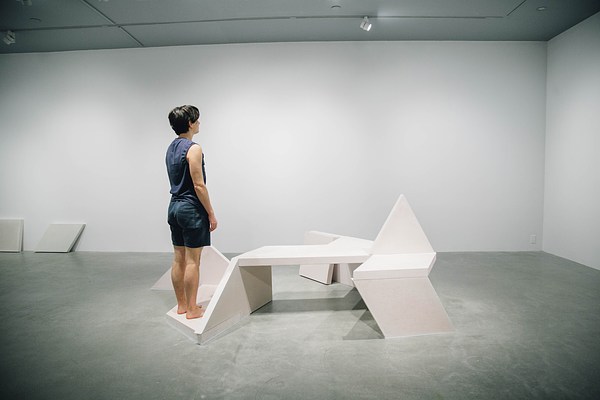 Performance piece where a barefoot person wearing shorts stands on the edge of a sculpture bench looking towards the back corner of the gallery away from the camera.&nbsp;