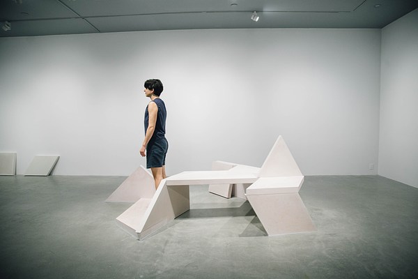Performance piece where a barefoot person wearing shorts stands on the edge of a sculpture bench looking towards the left wall of the gallery away from the camera.&nbsp;. 