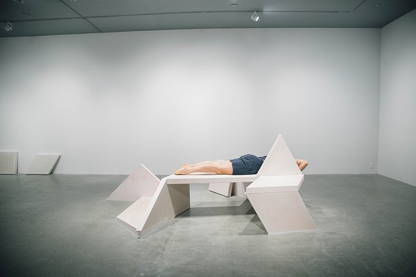 Performance piece where a barefoot person wearing shorts lies flat face down on a sculpture bench with their toes pointed.&nbsp;. 