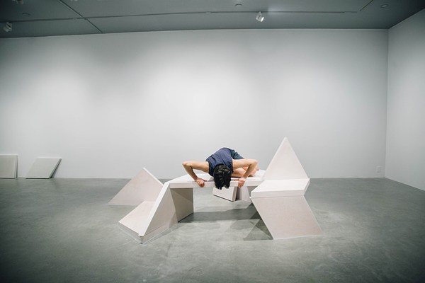 Performance piece where a barefoot person wearing shorts hunches over a sculpture bench with their arms supporting their upper body on the edge of the bench.&nbsp;. 
