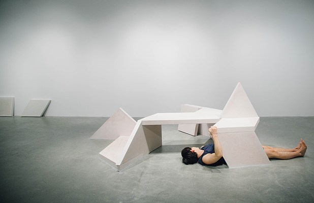 Performance piece where a barefoot person wearing shorts lies face up on the gallery wall partially underneath the sculpture bench with their hands holding onto the corner of the bench.&nbsp;. 