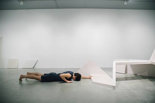 Performance piece where a barefoot person wearing shorts lies face down on the gallery floor with their head squished into the floor and an arm reaching out to touch the sculpture bench in front of them.&nbsp;. 