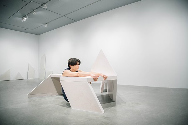Performance piece where a person squats next to a sculpture bench and holds onto the edge of the bench while leaning back.&nbsp;. 