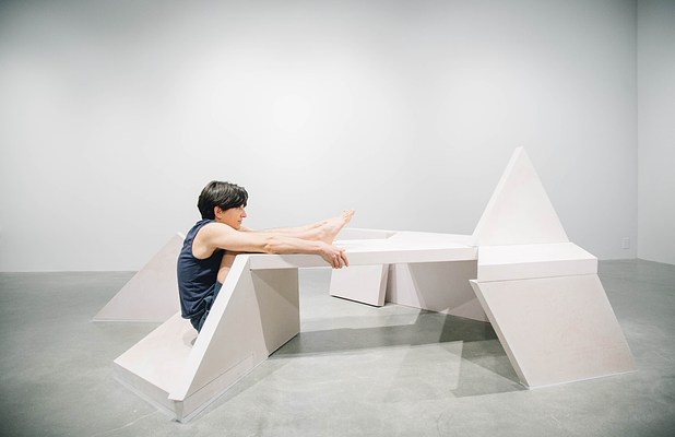 Performance piece where a person has their legs suspended on a sculpture bench and holds on with their arms while they sit on the floor.&nbsp;. 