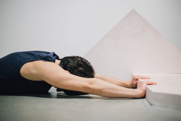 Close up performance piece where a person lies with their arms extended in front of a sculpture bench and grips the edge of the bench.&nbsp;. 