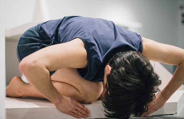 Performance piece where a person kneels over a sculpture bench with their head down and arms pressed into the edge of the bench.&nbsp;. 